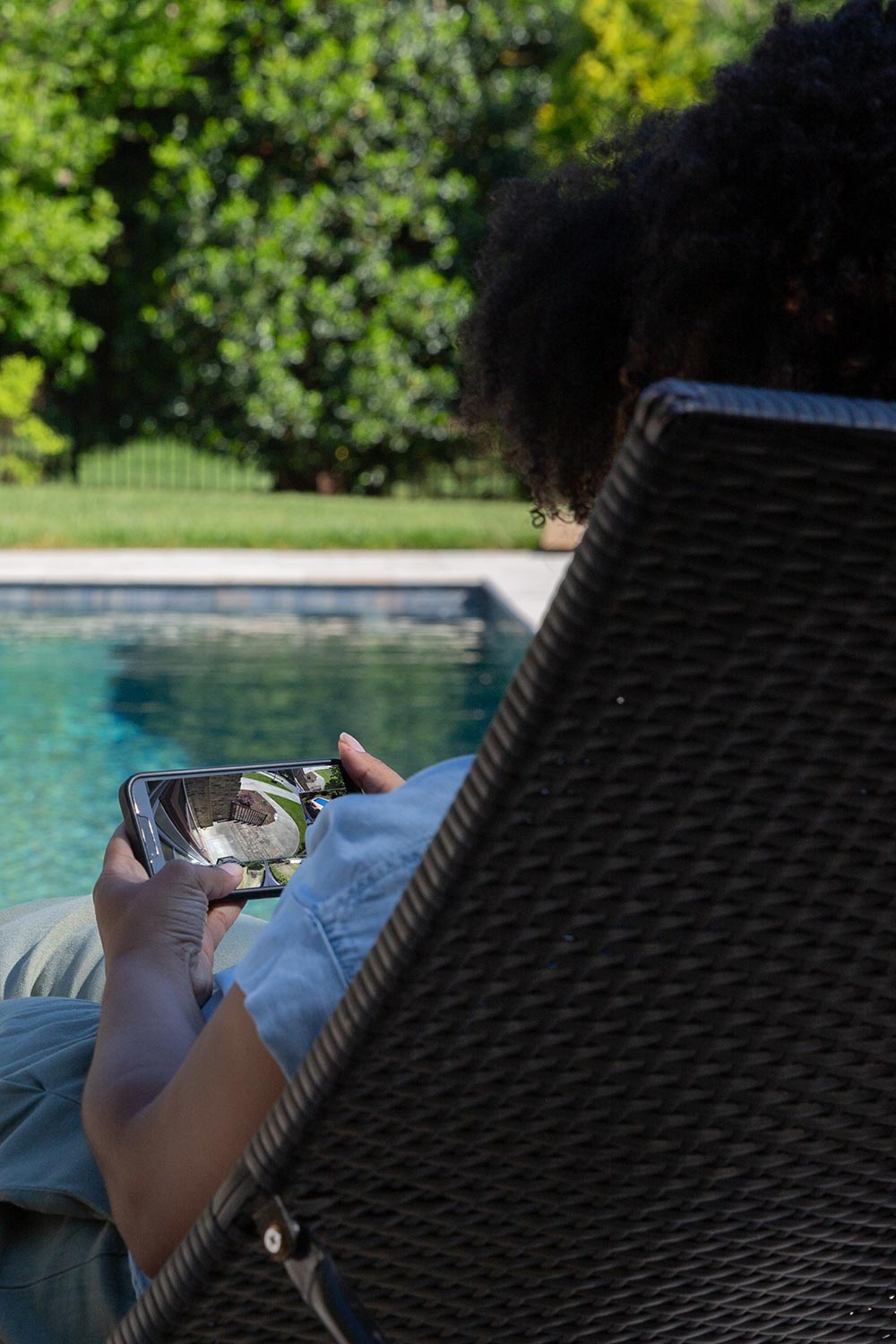 Person sitting by a pool checking a home security camera feed on a smartphone