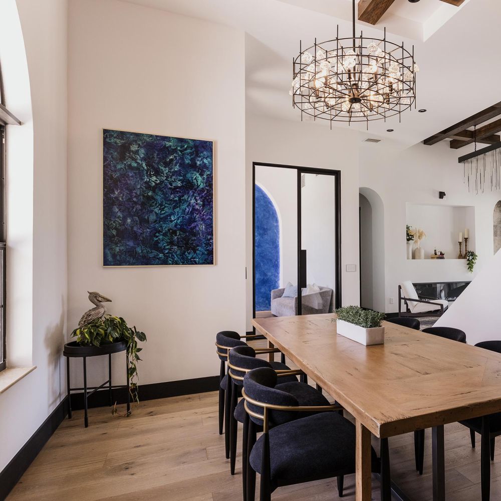 Wooden dining table with dark chairs beneath a wire chandelier next to abstract blue wall art